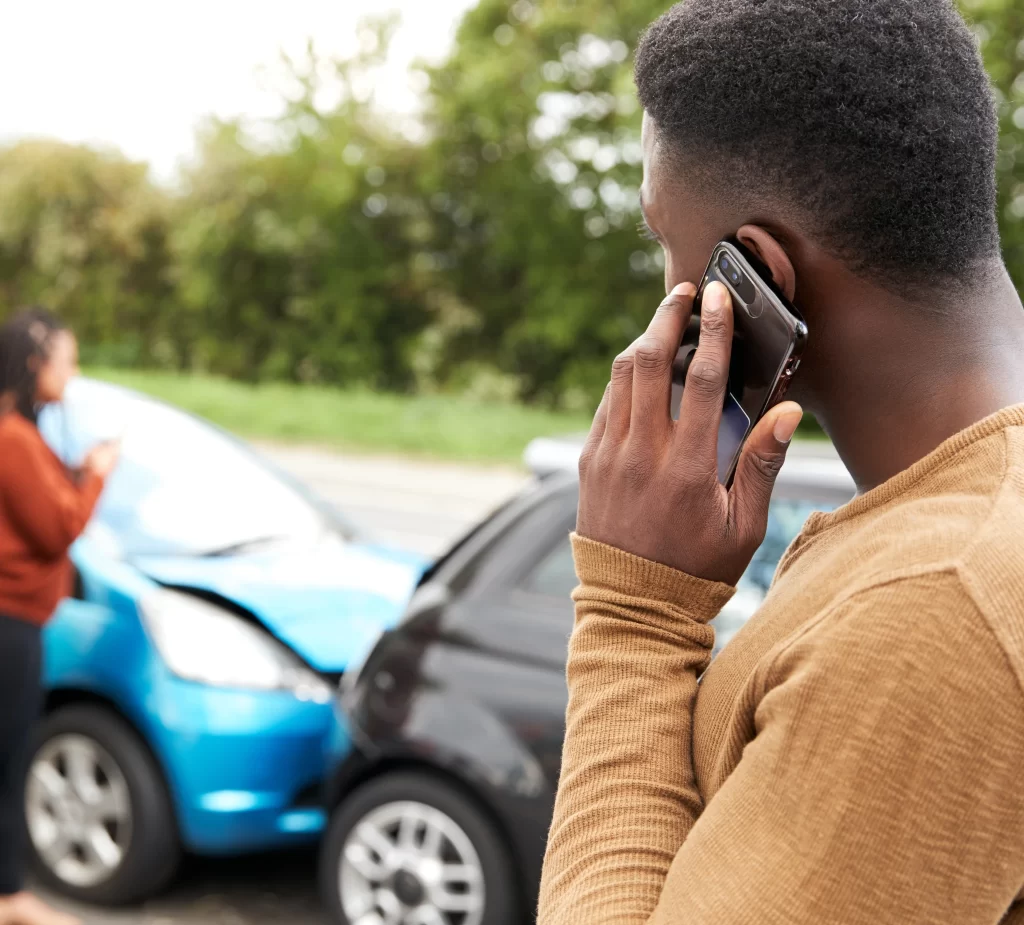 A man standing next to his car, holding a phone to report a center bender collision. Two vehicles with minor bumper damage are visible in the background, illustrating the process of filing an insurance claim.